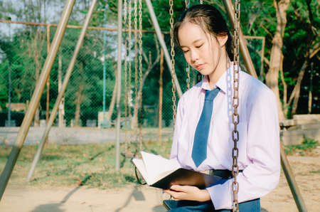 Lovely teenage girl wearing school uniform reading book while sitting on a swing at the park