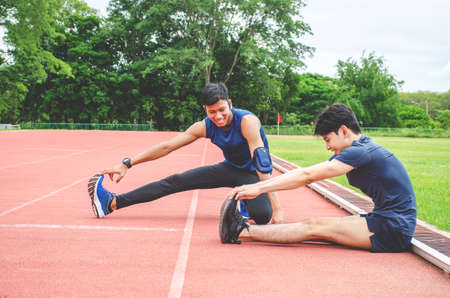 Training teammates. Two male athletes is warming-up,Two runners sitting on the racetrack at the stadium stretching their legs before runningの写真素材