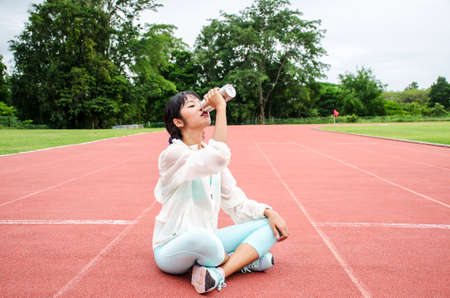 Young woman athlete sitting on runing track and drinking water in bottle.Cute slim young adult girl hold in hand bottle. Female sitting on running track and drinking bottled waterの写真素材