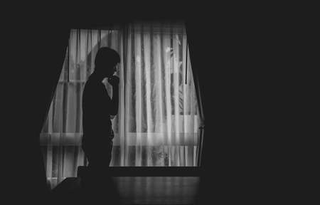 Alone man silhouette standing with hand on chin at the window closed with curtains in darkroom. Man stands at window alone, Depression and anxiety disorder concept.の写真素材