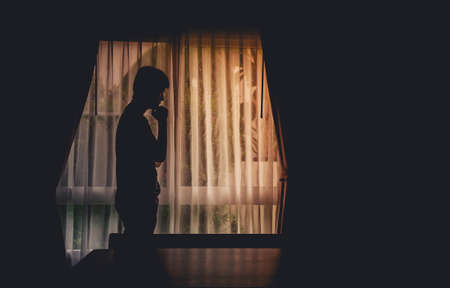 Alone man silhouette standing with hand on chin at the window closed with curtains in darkroom. Man stands at window alone, Depression and anxiety disorder concept.の写真素材
