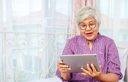 Portrait of smiling senior woman with eyeglasses using electronic tablet while relaxing at the bed...