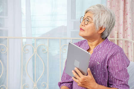 Portrait of smiling senior woman with eyeglasses thinking while looking up using electronic tablet...