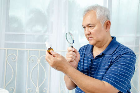 age, medicine, health care and people concept - Doubtful senior man looking at medicine in his hand with Magnifying glass at home or hospital officeの写真素材