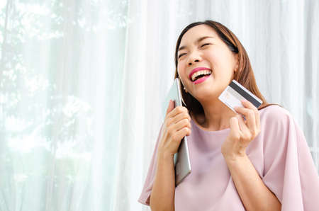 Smiling asian woman holding discount credit card in hand paying for shopping online at tablet in home, Young casual lifestyleの写真素材