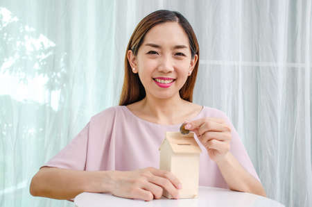 Asian woman putting money coin in to house piggy bank metaphor saving money financial for buy the homeの写真素材