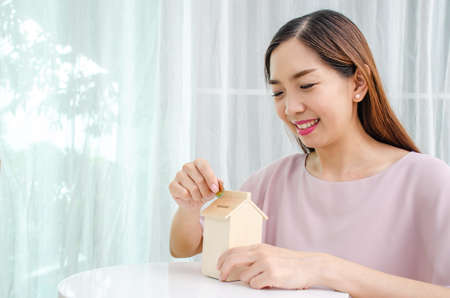 Asian woman putting money coin in to house piggy bank metaphor saving money financial for buy the homeの写真素材