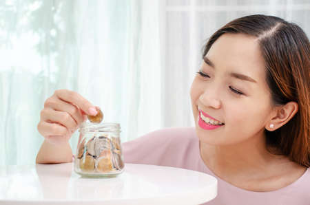 Young woman smile holding coins putting into glass jar, Savings concept for finance accounting at home or office.の写真素材