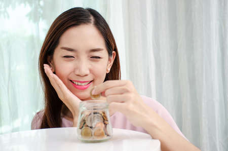 Young woman smile holding coins putting into glass jar, Savings concept for finance accounting at home or office.の写真素材