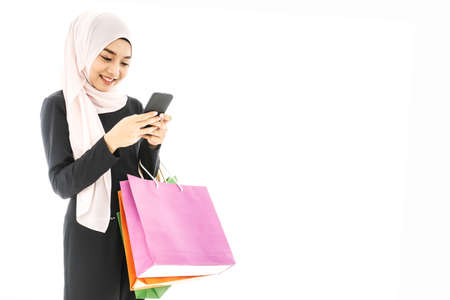 Portrait of beautiful asian muslim woman in a black hijab holding smartphone in one hand and paper bags after shopping day on white backgroundの写真素材