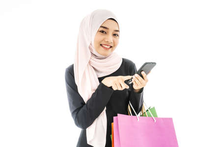 Portrait of beautiful asian muslim woman in a black hijab holding smartphone in one hand and paper bags after shopping day on white backgroundの写真素材