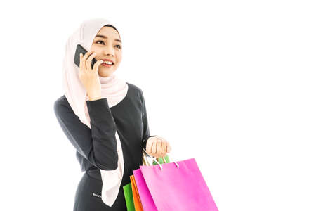 Portrait of beautiful asian muslim woman in a black hijab holding smartphone in one hand and paper bags after shopping day on white backgroundの写真素材