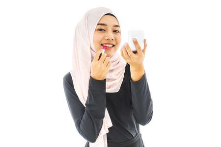 Portrait of smiley beautiful young muslim woman wearing Hijab and painting her lips with a lipstick with mirror on white background taken in studio. Using for Fashion and beauty conceptの写真素材