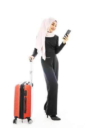 Portrait of beautiful asian muslim tourist traveller woman in a hijab holding red suitcase luggage get ready for traveling and trip and using cell phone isolate on white background.の写真素材