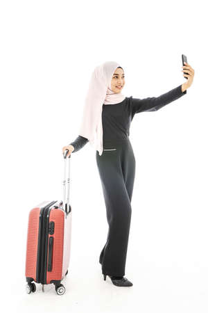 Portrait of beautiful asian muslim tourist traveller woman in a hijab holding red suitcase luggage get ready for traveling and trip and mobile phone making selfie isolate on white background.の写真素材