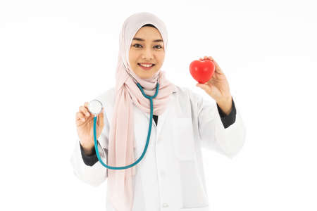 Portrait of beautiful young Muslim woman doctor shows a red heart on the left hand and wearing a stethoscope standing and smiles always happy and cares for the patients with joy isolated on white backgroundの写真素材