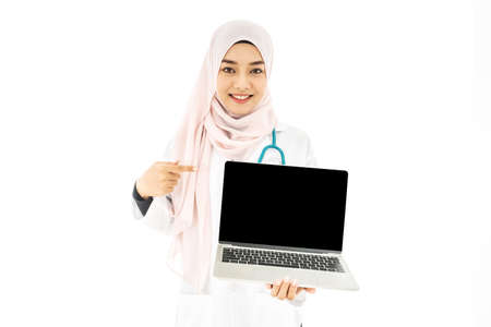Portrait of smiley beautiful young muslim woman doctor wearing doctor uniform and Hijab and Stethoscope holds laptop pointing with a finger at the white screen, standing isolated on white backgroundの写真素材