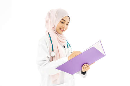 Portrait of smiley beautiful young muslim woman doctor wearing doctor uniform and Hijab and Stethoscope writing in her note book, standing isolated on white backgroundの写真素材