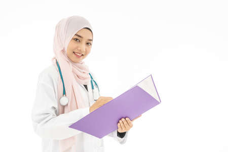 Portrait of smiley beautiful young muslim woman doctor wearing doctor uniform and Hijab and Stethoscope writing in her note book, standing isolated on white backgroundの写真素材