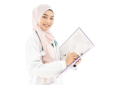 Portrait of smiley beautiful young muslim woman doctor wearing doctor uniform and Hijab and Stethoscope writing in her note book, standing isolated on white backgroundの写真素材