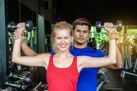 sport, fitness, lifestyle and people concept, The girl is training dumbbells flexing muscles with help of her personal trainer in the gym.の写真素材