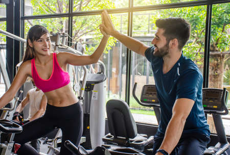 Young sporty woman and man giving each other a high five after cycling training in gym. Fit couple high five after workout in health club, Fitness class doing sport biking in the gym for health.の写真素材