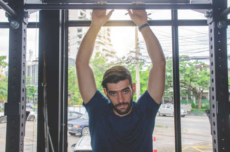 fitness, sport, training and lifestyle concept - Portrait of handsome strong athletic man exercising on horizontal bar in fitness gym and looking at cameraの写真素材