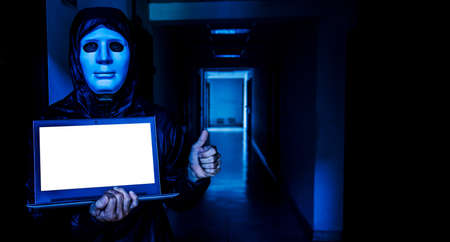 Anonymous computer hacker in white mask and hoodie. Obscured dark face holds a laptop tight in his hands on target room background, Data thief, internet attack, darknet and cyber security conceptの写真素材