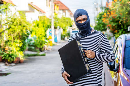 The male thief wears a black and white striped shirt and wears a black mask, smiling happily after stealing briefcase with money he wants.の写真素材