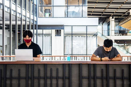 Two asian man wear a face mask for protecting the virus while working in office with the laptop in casual and keep social distancing. New normal concept.の写真素材