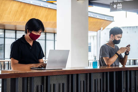 Two asian man wear a face mask for protecting the virus while working in office with the laptop in casual and keep social distancing. New normal concept.の写真素材