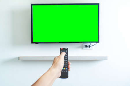 Hand holding TV remote controller over green screen a modern television set. Place for your advertisement.の写真素材