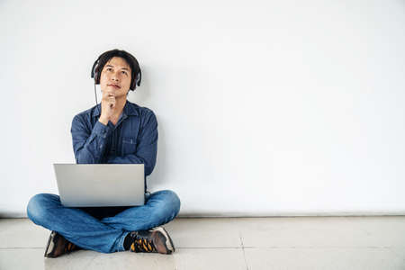Young man asian sitting on the floor with using computer laptop and listening data in feel doubting and confused or thinking about an idea posing isolated over white wall,  copy spaceの写真素材