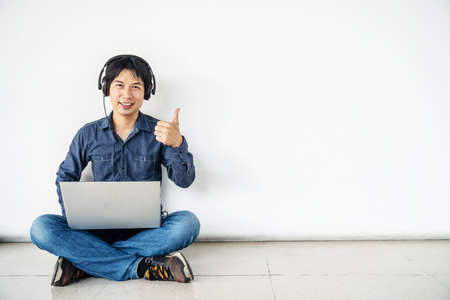 Young man asian sitting on the floor with using computer laptop listening nice music is happy with big smile doing ok sign, thumb up with fingers, excellent sign, copy spaceの写真素材