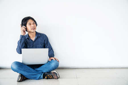 Young man asian sitting on the floor with using computer laptop smiling and listening nice music and feel relieved from stressed posing isolated over white wall, copy spaceの写真素材
