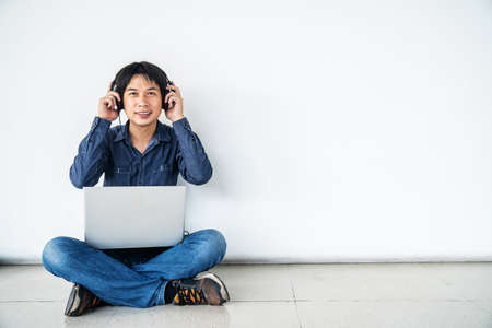 Young man asian sitting on the floor with using computer laptop smiling and listening nice music and feel relieved from stressed posing isolated over white wall, copy spaceの写真素材