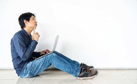 Young man asian sitting on the floor with using computer laptop doubting and confused or thinking about an idea posing isolated over white wallの写真素材