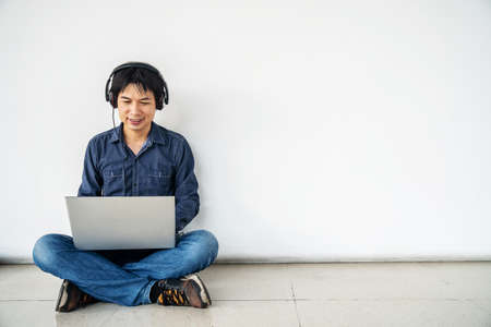 Young man asian sitting on the floor with using computer laptop smiling and listening nice music and feel relieved from stressed posing isolated over white wall, copy spaceの写真素材