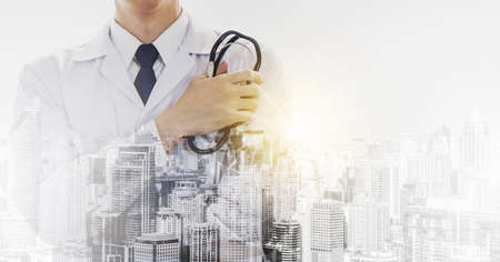 Front view of smart young male doctor crossed arm and holding stethoscope on white sterile coat while standing outdoors with abstract city view on background. Medical industry. Double exposureの写真素材