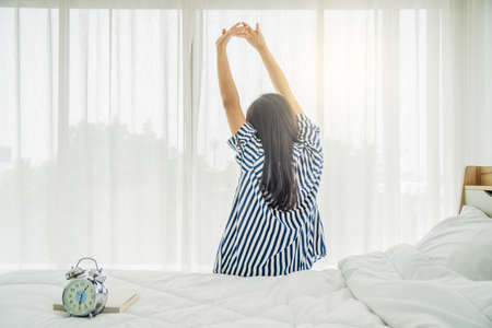 Back view of Beautiful young woman sitting on her bed in morning, stretching with arms raised, There is an alarm clock that tells the time Healthy lifestyle, Wellness concept, free spaceの写真素材