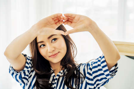 Beautiful asian young woman sitting on bed and making heart shape with fingers. Portrait of smiling lady posing in her bedroom, Concept happiness and love.の写真素材