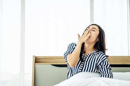 rest, holidays, comfort and people concept, Portrait of sleepy asian woman in sleepwear yawning after her waking up so early in the morning, Sitting on bedの写真素材
