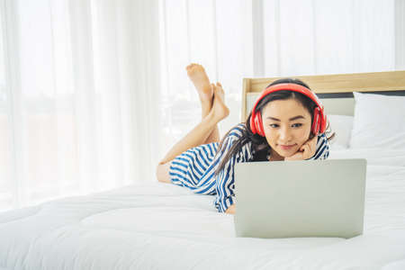 Beautiful young woman relaxing and listening the music while using laptop computer on the bed in bed room with copy space. Beauty asian girl gets happy, relaxing. She enjoying lifeの写真素材