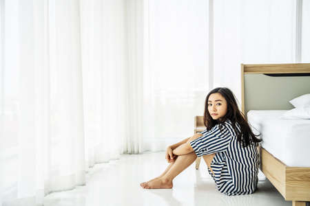 Sad asian woman lonely at home self isolation quarantine for COVID-19 Coronavirus social distancing prevention, She sitting on the floor near bed feeling boring in bedroom, Mental health.の写真素材