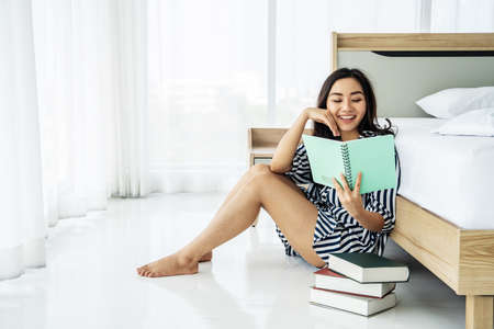 Happy beautiful woman wearing sleepwear reading a book and sitting near bed in morning. Resting and Relaxation time.の写真素材