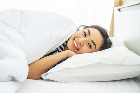 Young beautiful woman smiling while lying on white pillow in bed at early morningの写真素材