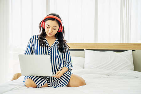 quarantine due coronavirus pandemic. stay at home. beautiful young woman relaxing and listening the music while using laptop computer on the bed in bed room. Beauty asian girl gets happy, relaxing.の写真素材