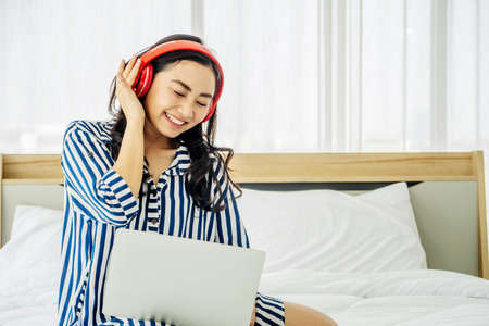quarantine due coronavirus pandemic. stay at home. beautiful young woman relaxing and listening the music while using laptop computer on the bed in bed room. Beauty asian girl gets happy, relaxing.の写真素材