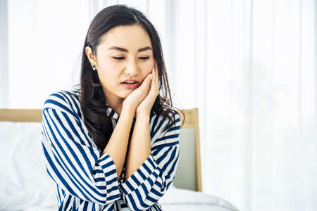 Young Asian woman touching cheek while suffering from toothache and discomfort on bed in white bedroom morning.Concept of women's health care.の写真素材