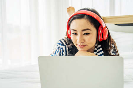 quarantine due coronavirus pandemic. stay at home. beautiful young woman relaxing and listening the music while using laptop computer on the bed in bed room. Beauty asian girl gets happy, relaxing.の写真素材